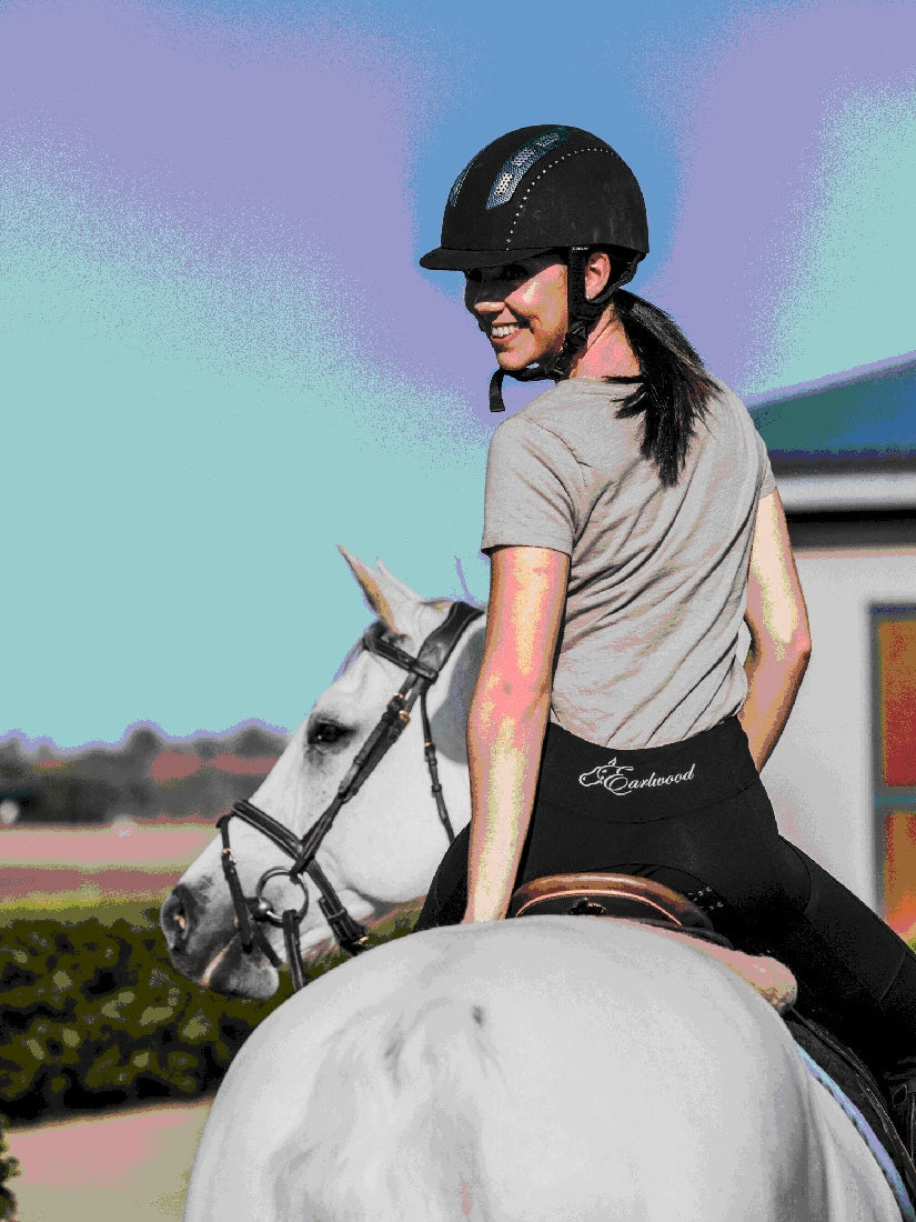 Woman smiles wearing helmet and horse riding tights on white horse.