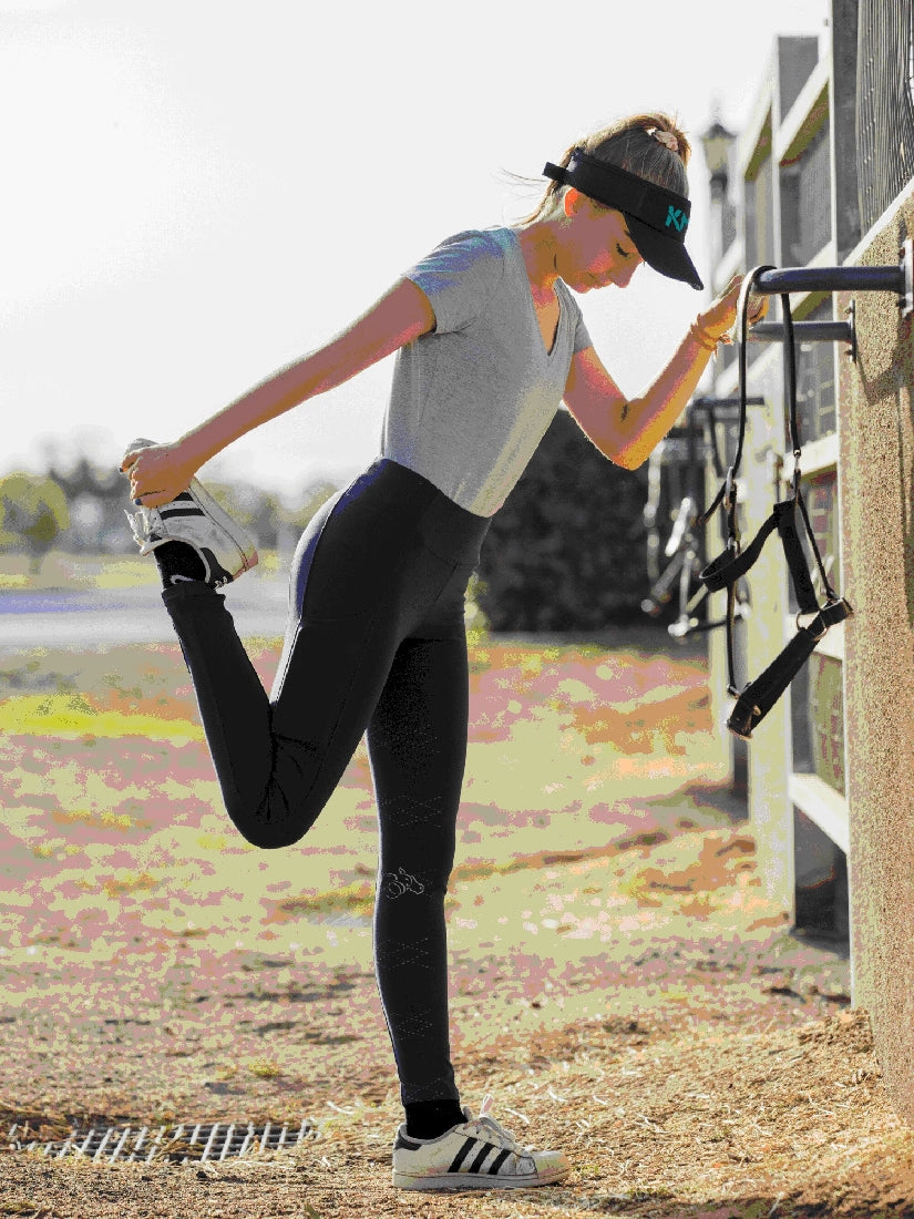 Woman stretching leg, wearing visor and black horse riding tights.